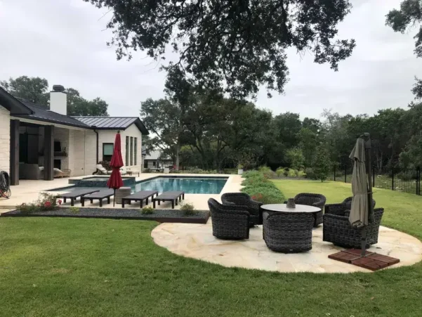 Luxury backyard landscape design in Bee Cave, TX, featuring an in-ground swimming pool, spa, custom circular flagstone patio with outdoor seating, and a manicured green lawn under oak trees.